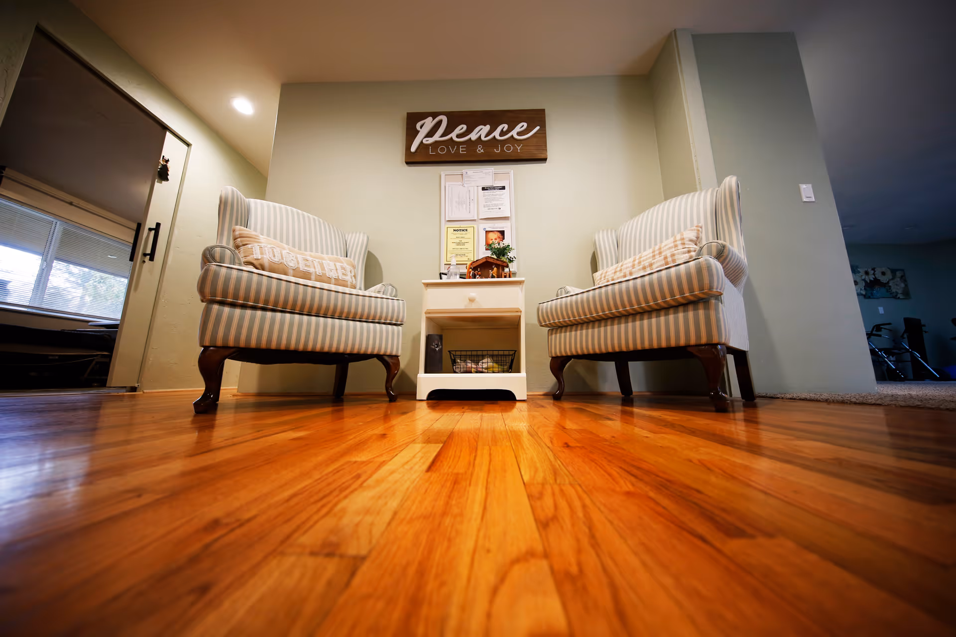 Two striped armchairs with pillows that say 'TOGETHER' facing each other with a small white side table between them. Above the table is a wall sign that reads 'Peace LOVE & JOY'. The room has wooden flooring and light-colored walls.