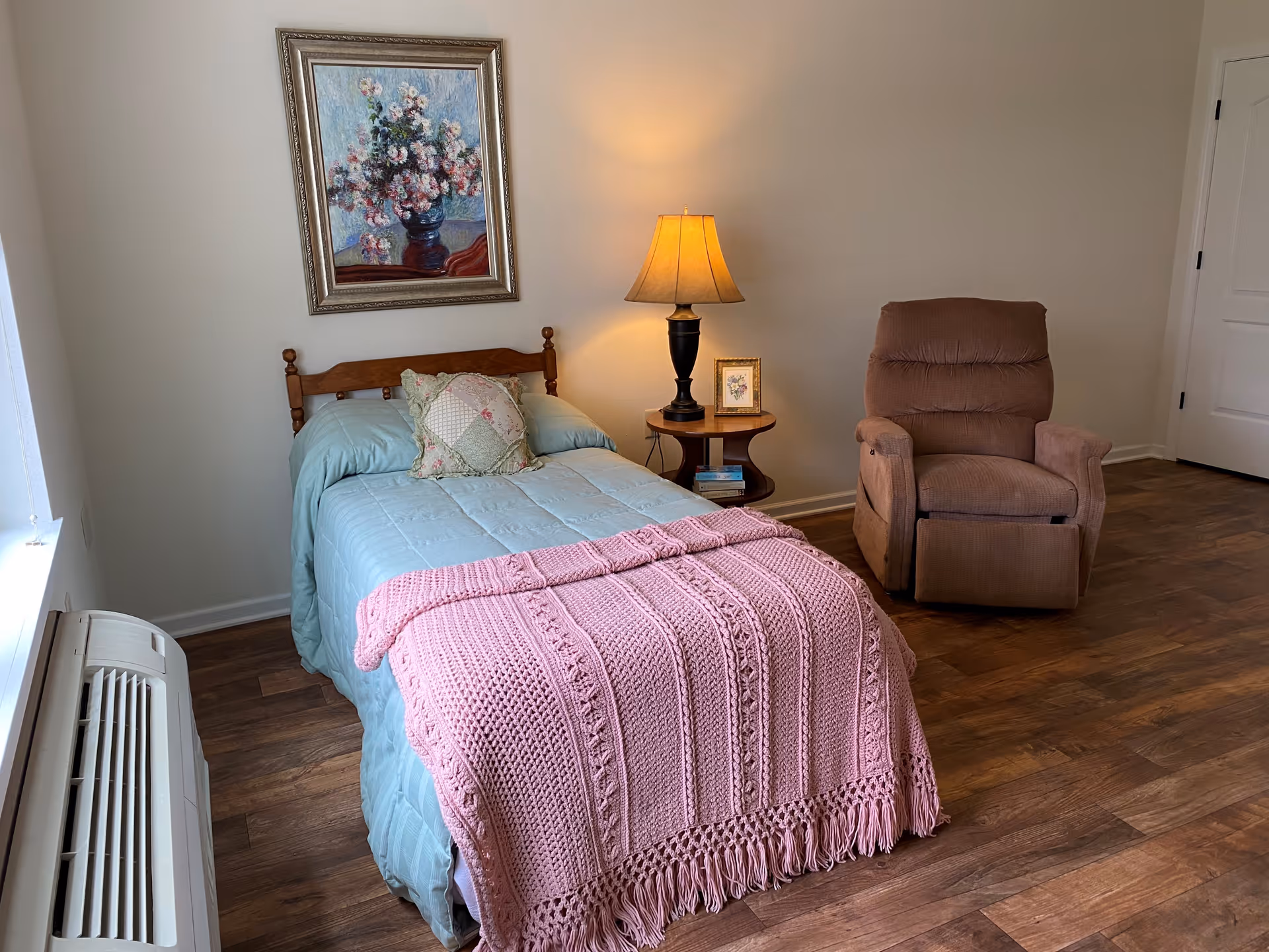 A cozy bedroom with a single bed covered in a light blue comforter and a pink knitted blanket at the foot. There is a decorative pillow on the bed. Next to the bed is a wooden side table with a lamp, a small framed picture, and some books. A brown recliner chair is positioned near the table. The room has wooden flooring, a window with an air conditioning unit below it, and a framed floral painting hanging on the wall above the bed.