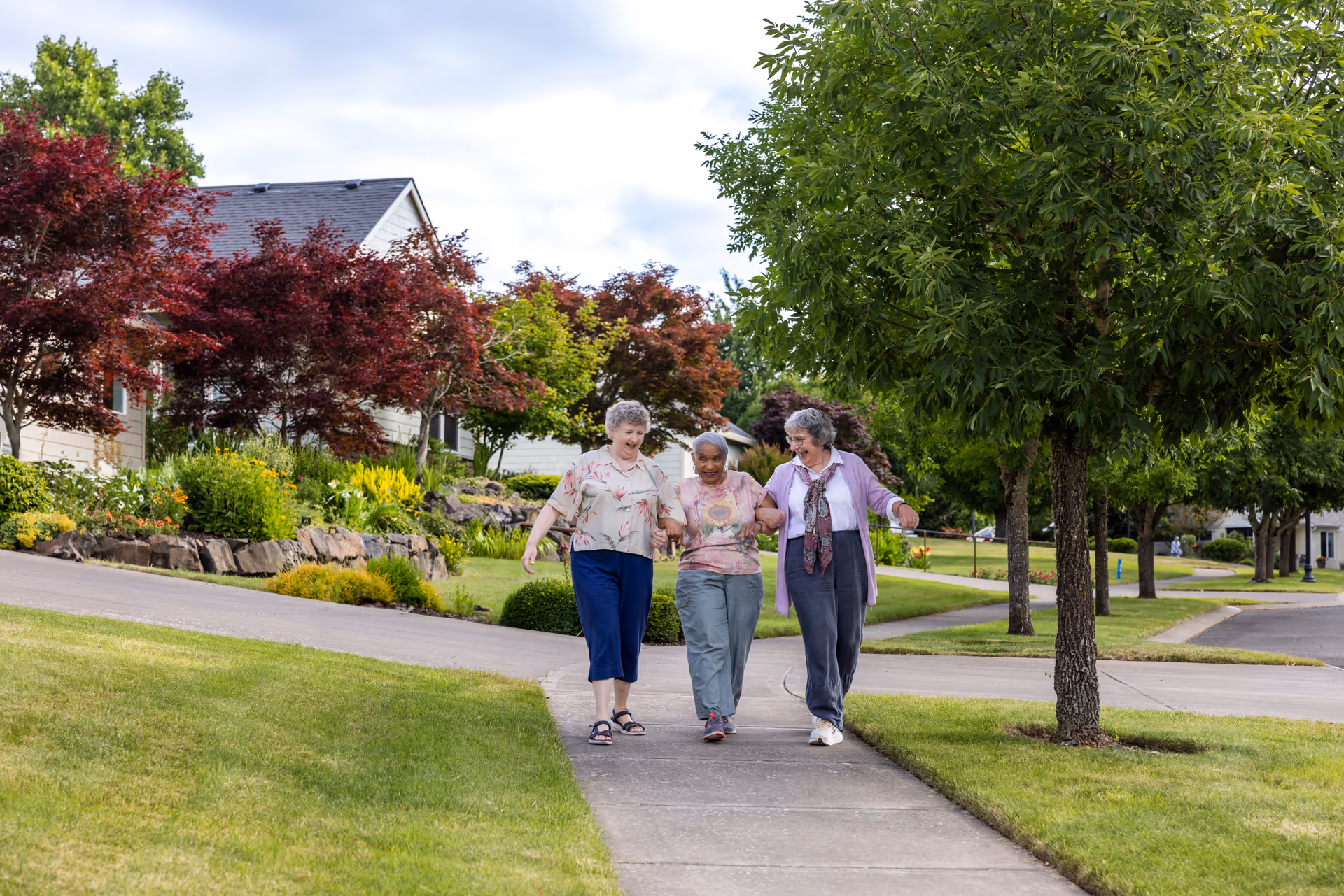 Three elderly women walking arm in arm on a sidewalk in a residential neighborhood with green lawns, trees, and houses in the background under a partly cloudy sky.