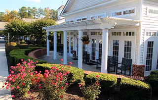 White pergola-style covered porch with chairs at the front of a light-colored senior living building, surrounded by manicured hedges and pink flowering shrubs.