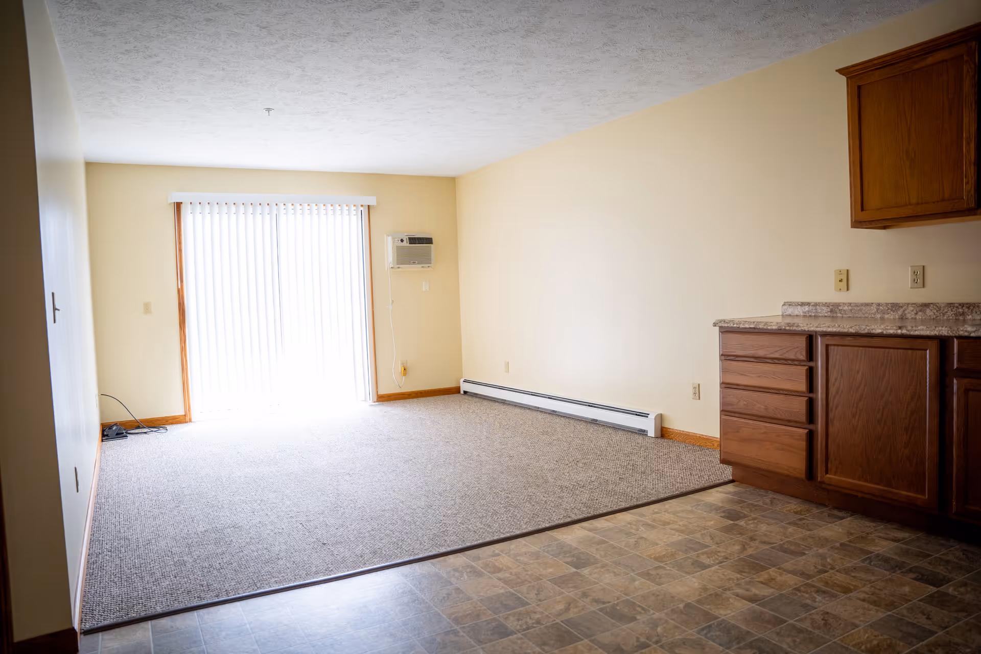 Empty room with beige walls and carpeted floor area next to a tiled floor section with wooden cabinets and countertop. A large sliding glass door with vertical blinds allows natural light into the room. A wall-mounted air conditioning unit is visible next to the door.