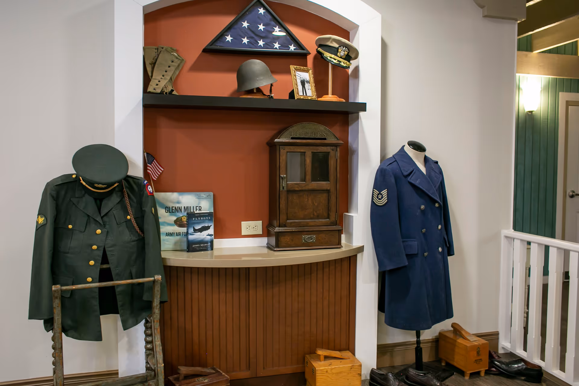 A display area featuring vintage military uniforms on mannequins, a folded American flag in a triangular case, military helmets, a framed photograph, and books about Glenn Miller and Army Air Force Flyboys. The display is set against a red wall with a wooden counter and various small wooden boxes and shoes on the floor.