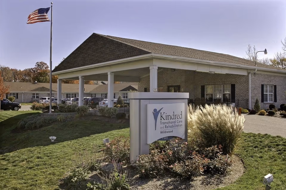 Exterior view of Kindred Transitional Care and Rehabilitation Wildwood building with a covered entrance, an American flag on a flagpole, and landscaped greenery in front.