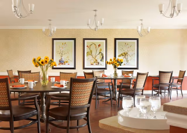 A dining room with several round tables set with white mugs, orange napkins, and vases of sunflowers. The room has wooden chairs with patterned backs, three framed botanical prints on the wall, and multiple chandeliers hanging from the ceiling. In the foreground, there is a tray with glasses of water on a counter.