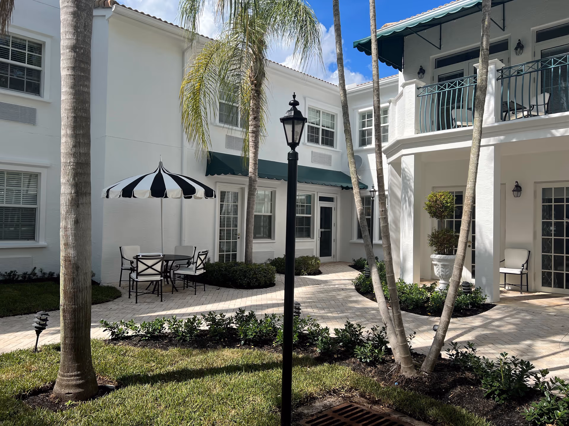 Outdoor courtyard area of a senior living facility with white buildings, palm trees, a black and white striped umbrella over a table with four chairs, green awnings over windows and doors, and a black lamppost in the center.