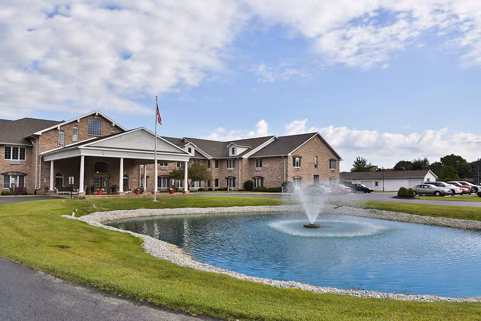 Brick senior living facility with a covered entrance, flagpole, and a pond with a fountain in the foreground.