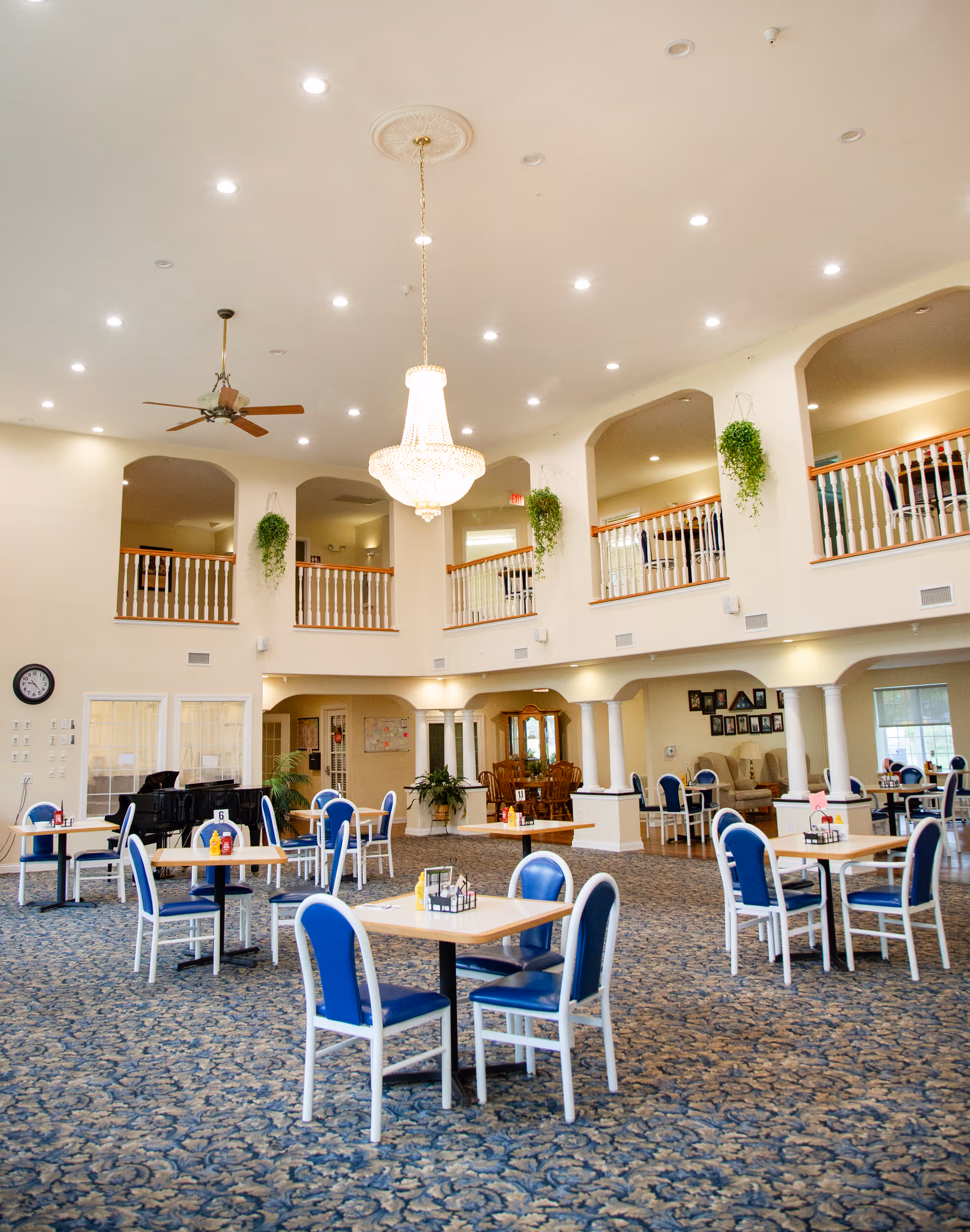 Spacious dining area in an assisted living facility with multiple tables and blue cushioned chairs arranged on a patterned carpet. The room features a high ceiling with recessed lighting, a large chandelier, ceiling fan, and hanging plants. There is a second-floor balcony with white railings overlooking the dining area, and a piano is visible in the corner.