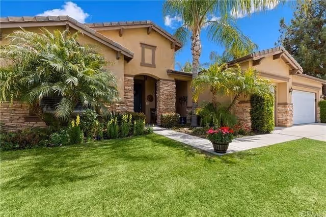 Exterior view of a single-story residential building with a well-maintained green lawn, palm trees, and various shrubs. The building features a stone and stucco facade with a covered entrance and a two-car garage under a clear blue sky.