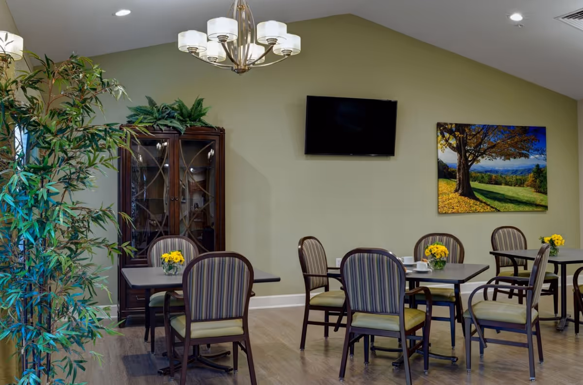 Dining area with several tables and striped chairs, a wall-mounted TV, a cabinet, and framed landscape artwork.