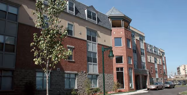 Front exterior of a three-story brick and stucco retirement residence with dormer windows, a turret-like entrance, street lamps, and parked cars.