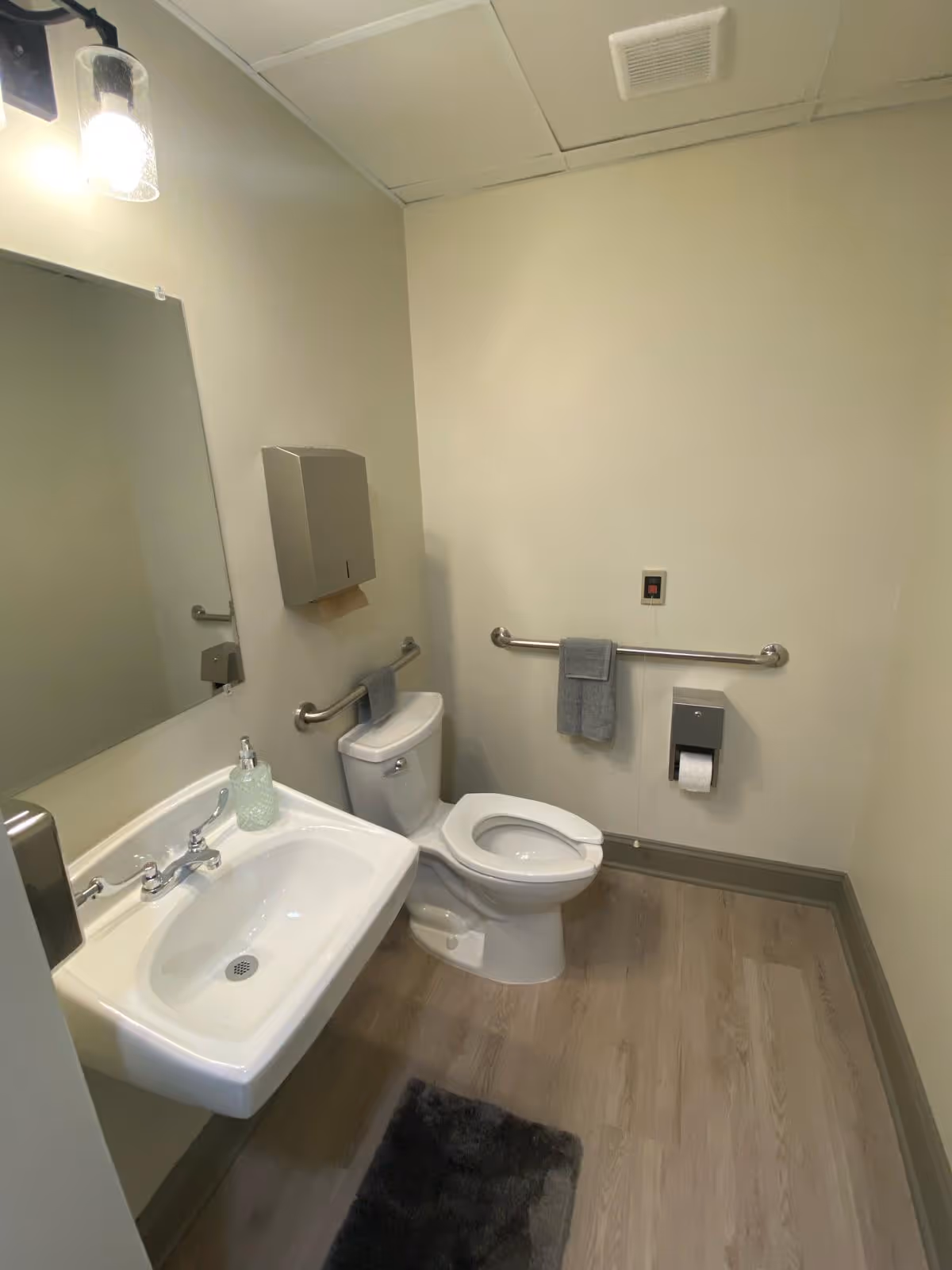 A clean and accessible bathroom with a white sink, a mirror above it, a toilet with a raised seat, grab bars on the walls, a paper towel dispenser, a soap dispenser, and a gray towel hanging on one of the grab bars. The floor has light wood-style flooring and a dark gray rug in front of the sink.