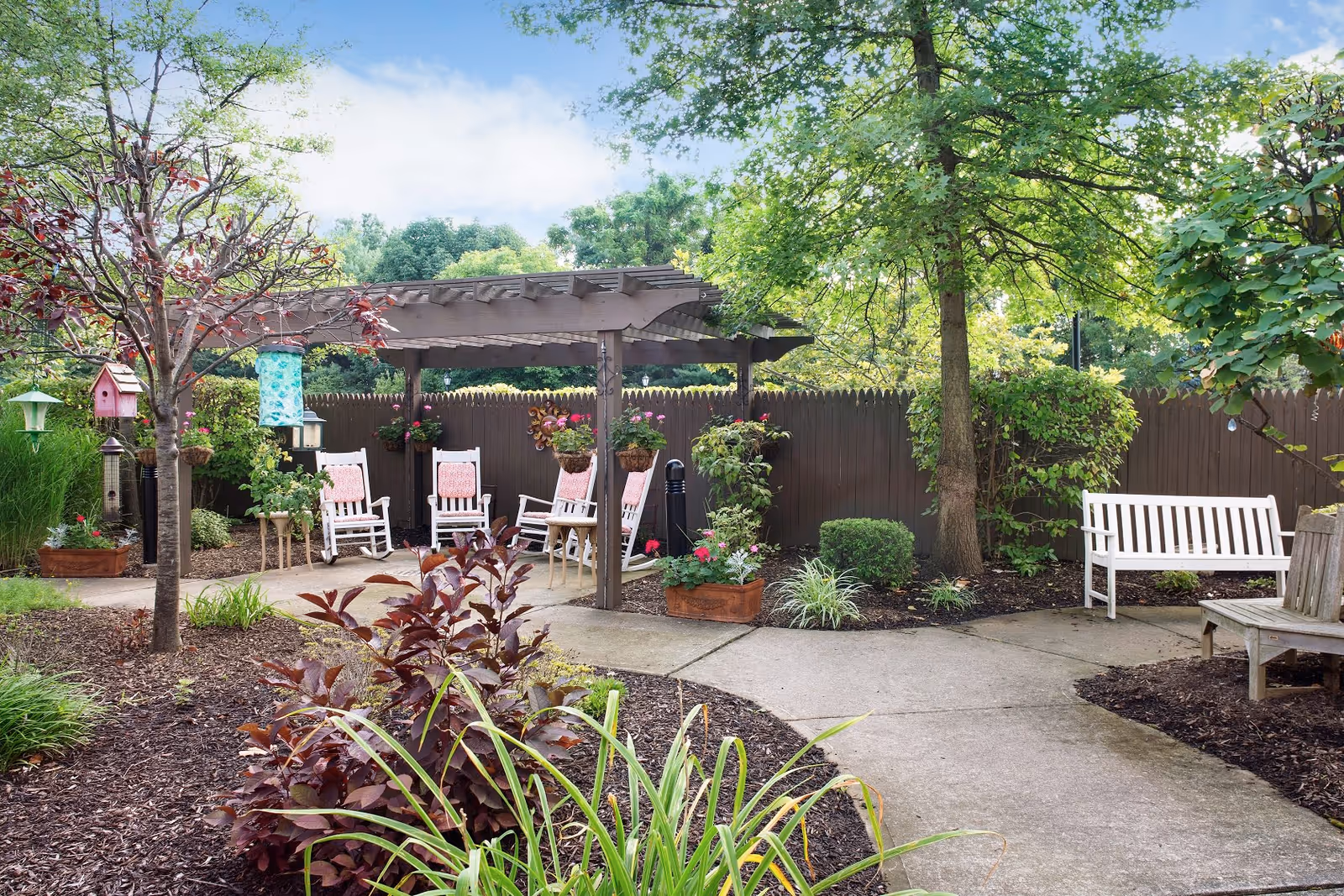 A peaceful outdoor garden area with a wooden pergola covering several white rocking chairs with pink cushions. The garden is surrounded by a wooden fence and features various plants, trees, hanging flower pots, birdhouses, and benches along a paved walkway.