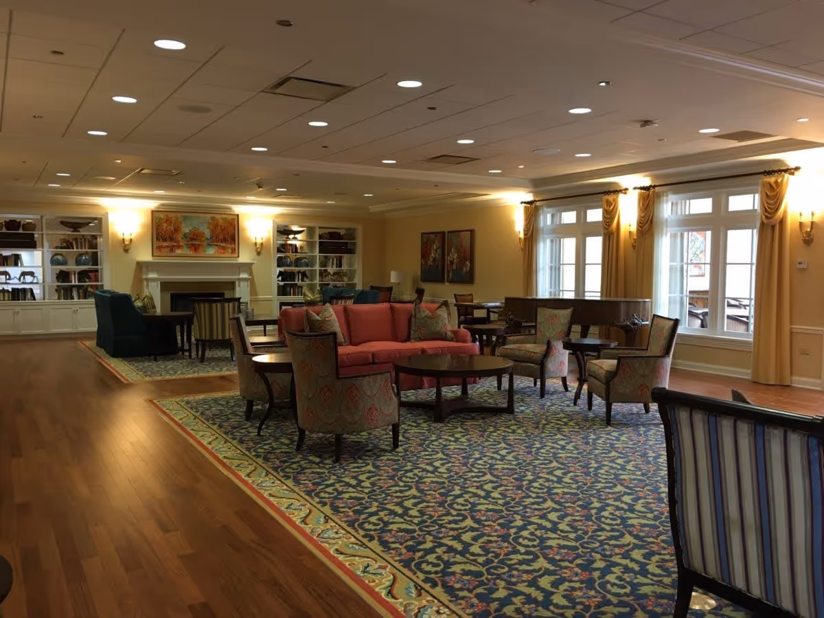 A spacious and well-lit living room area with a large patterned carpet, multiple upholstered chairs, a red sofa, round wooden tables, built-in bookshelves, a fireplace, and large windows with yellow curtains allowing natural light to enter.