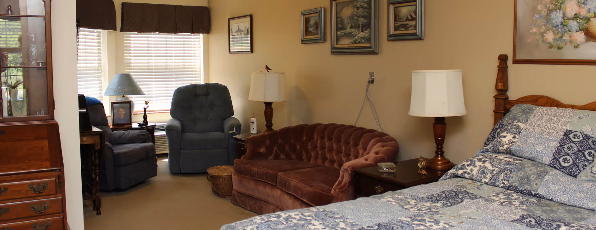 A cozy room with a bed covered in a blue and white patterned quilt, a brown tufted loveseat, two blue recliner chairs near a window with blinds, wooden side tables with lamps, and framed artwork on the walls.