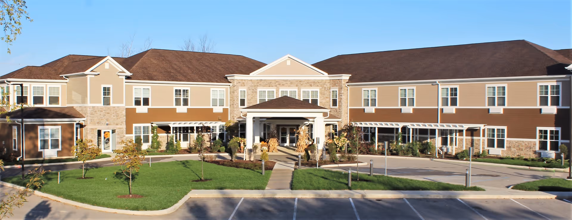 Front exterior view of a two-story assisted living facility building with a brown roof, beige and brown siding, and stone accents. The entrance has a covered porch with columns and seasonal decorations. There is a well-maintained lawn with small trees and a parking area in front under a clear blue sky.