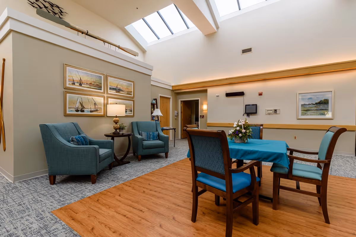 A bright and cozy common area in a senior living facility with two teal armchairs and a small round table with a lamp and plant between them. There is a wooden table covered with a blue tablecloth and four chairs around it. The room has a high ceiling with skylights, framed paintings on the walls, and a wooden floor section under the table.