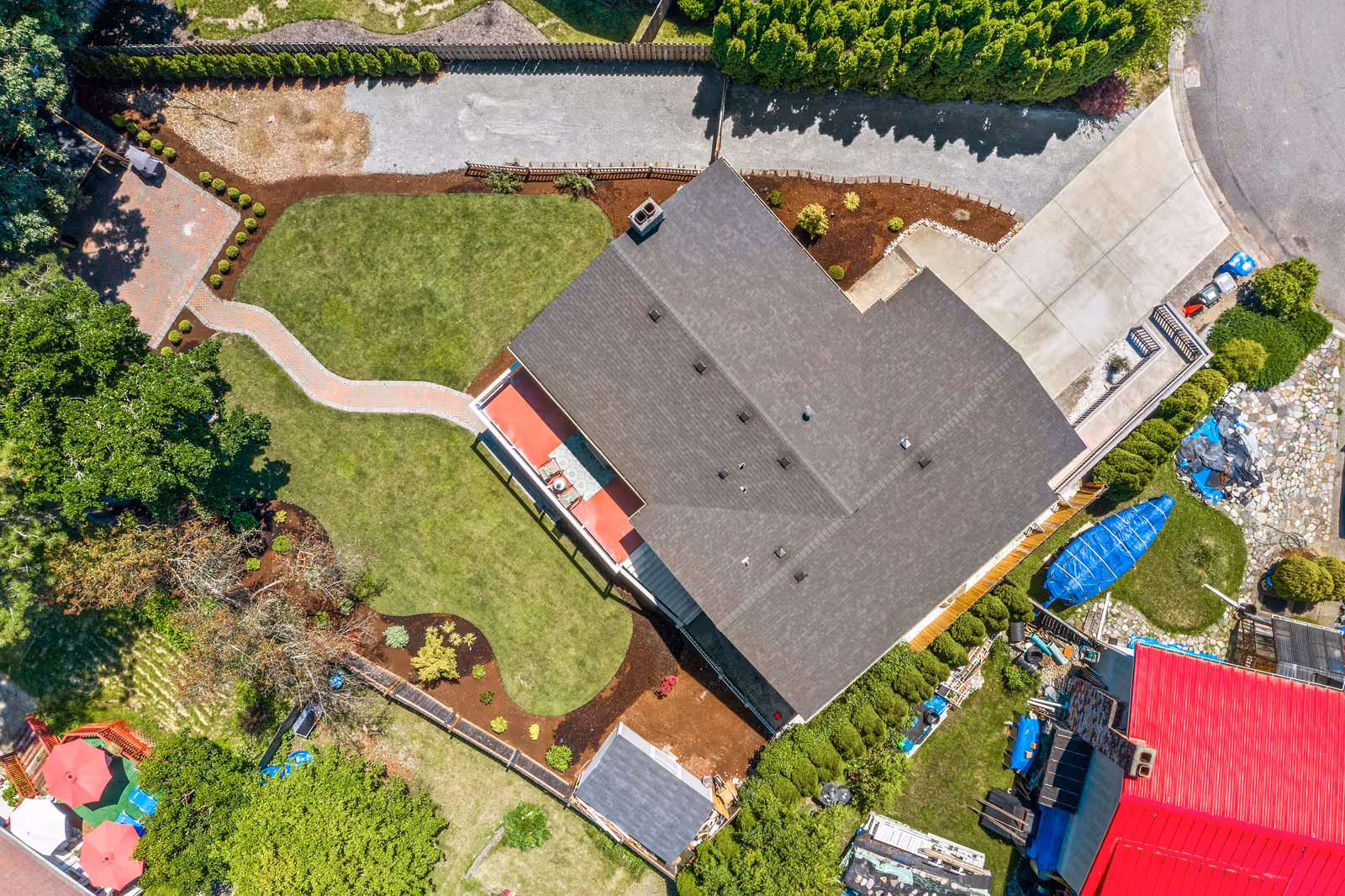 Aerial view of a residential property with a large house surrounded by a well-maintained lawn, garden beds, paved walkways, and various trees and shrubs. The driveway and neighboring properties are also visible.