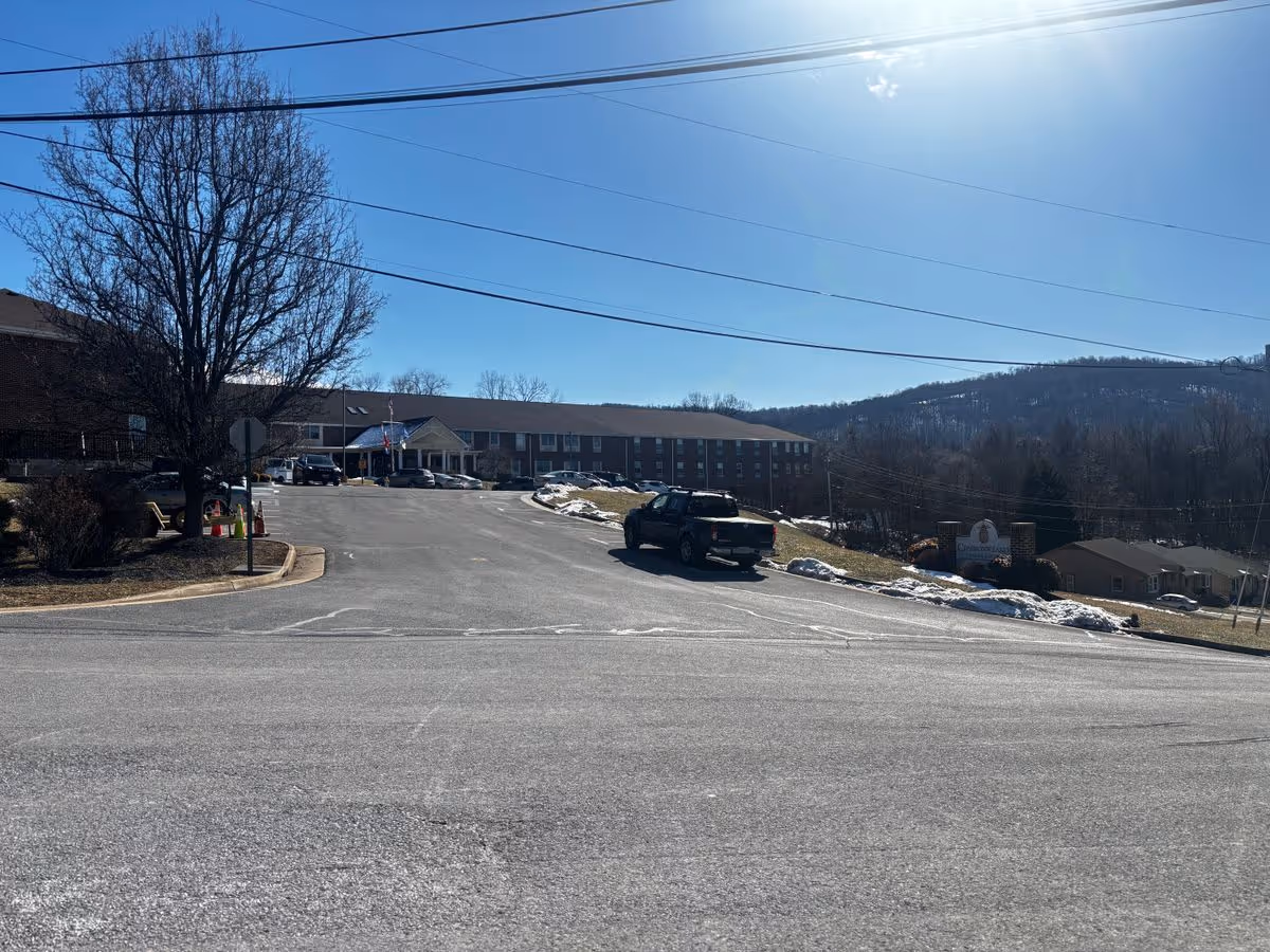 Front exterior of a long brick senior living building with a driveway, parked truck, entrance canopy and hills in the background.