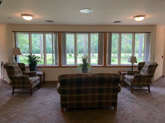 A cozy sitting area with a plaid sofa and two matching armchairs arranged around a window with a view of green trees and grass. There are two side tables with lamps and a plant on the windowsill.