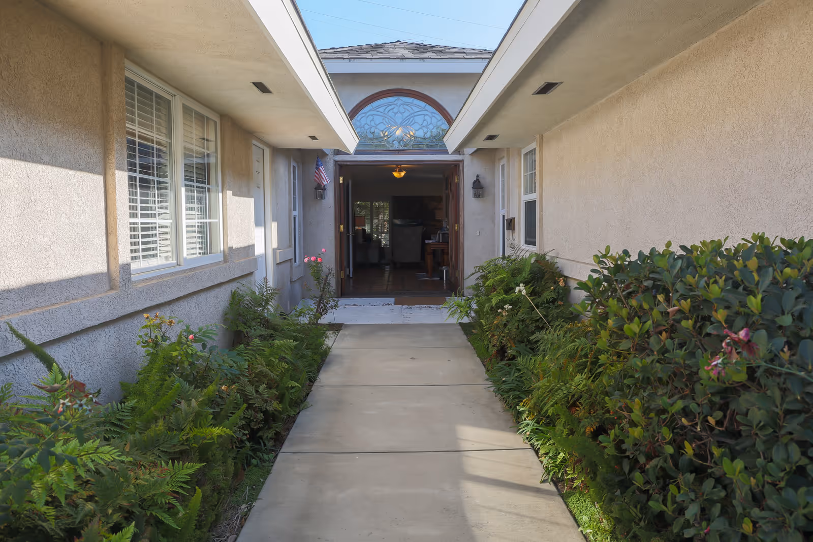 A concrete walkway leading to the entrance of a building with beige stucco walls on both sides. There are green plants and bushes lining the walkway. The entrance has a decorative glass window above the door and the door is open, showing a glimpse of the interior with furniture and lighting.
