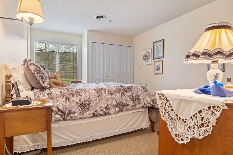 A cozy bedroom with a bed covered in a floral patterned comforter and pillows, a wooden nightstand with a lamp and clock, a window with blinds showing trees outside, a closet with double doors, and framed pictures on the wall. A wooden dresser with a lace doily and a lamp with a decorative shade is also visible.