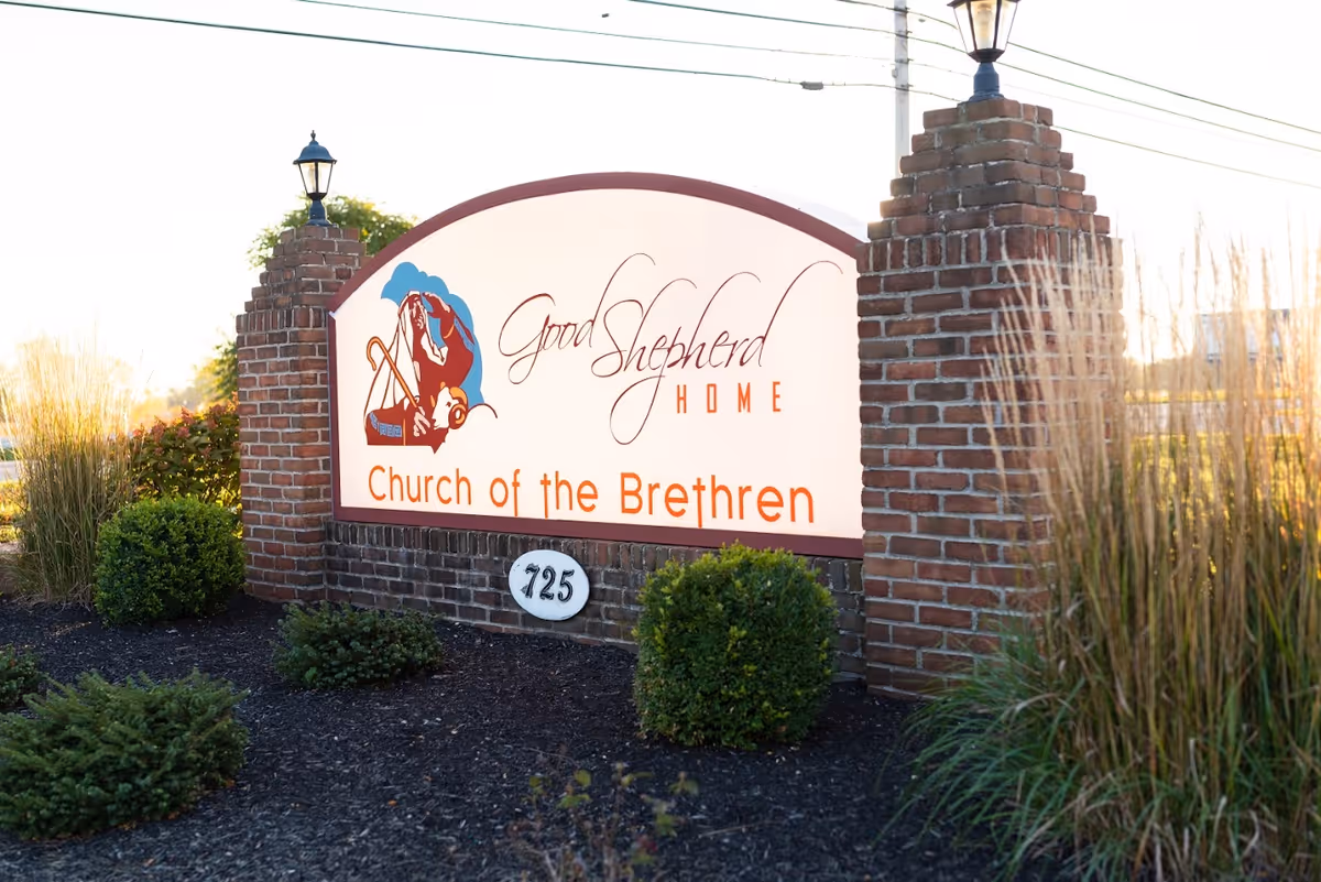 Outdoor sign for Good Shepherd Home Church of the Brethren, mounted on a brick structure with two lamp posts on top, surrounded by landscaping with bushes and tall grasses.