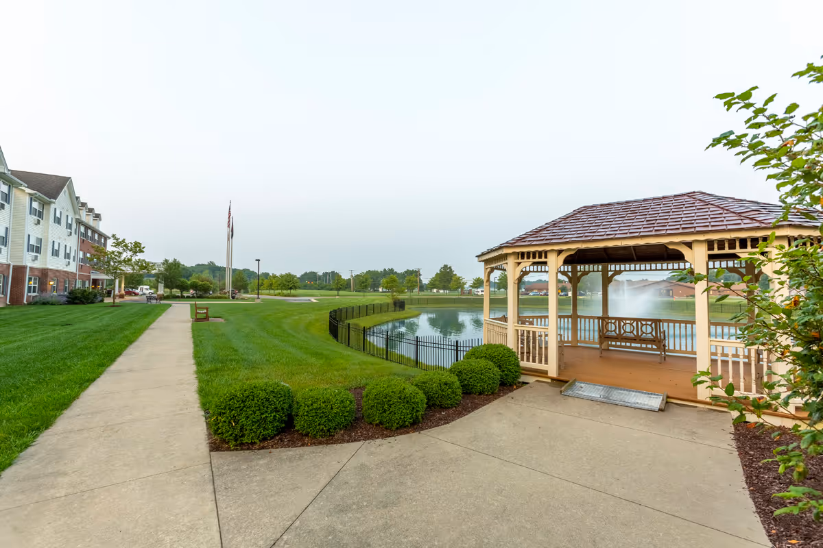 A gazebo beside a pond with a walking path, manicured lawn, and the senior living building in the background.