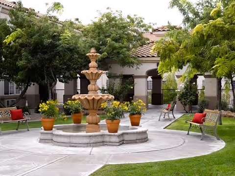 Outdoor courtyard area with a multi-tiered stone fountain in the center, surrounded by potted yellow flowers. There are benches with red cushions placed around the fountain, and trees and greenery provide shade. In the background, there is a building with arched walkways and a tiled roof.