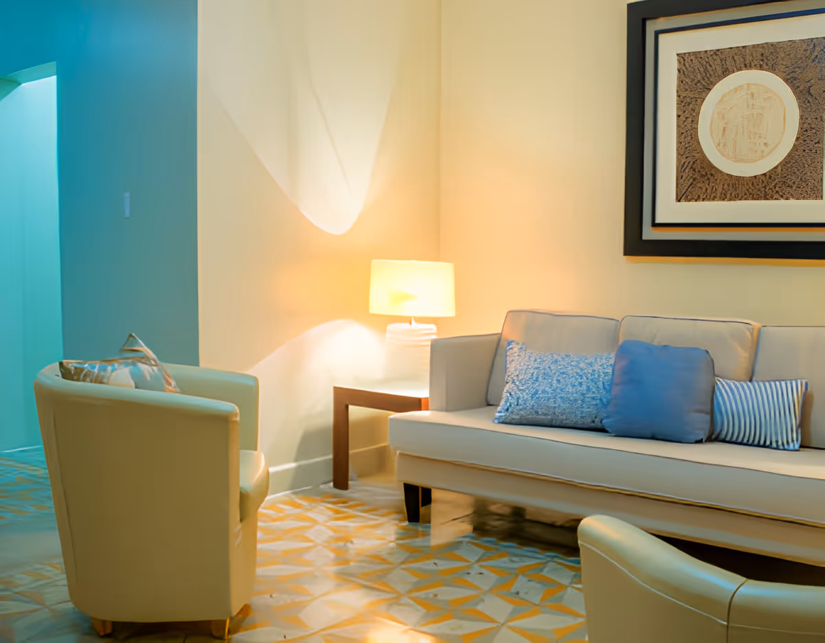 A cozy living room area with a beige sofa adorned with three decorative pillows in blue and striped patterns, two beige armchairs with patterned cushions, a wooden side table with a lit table lamp, and a framed abstract artwork on the wall.