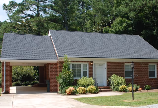 Single-story brick building with a dark shingled roof, white door, and windows surrounded by green shrubs and trees. There is a driveway and a covered carport on the left side, and a black lamp post on the right side near the lawn.