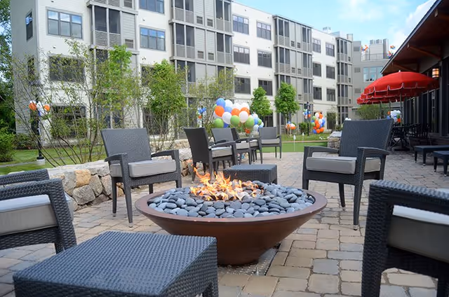 Outdoor courtyard with a central stone fire pit surrounded by wicker chairs and tables, colorful balloons and a multi-story building in the background.