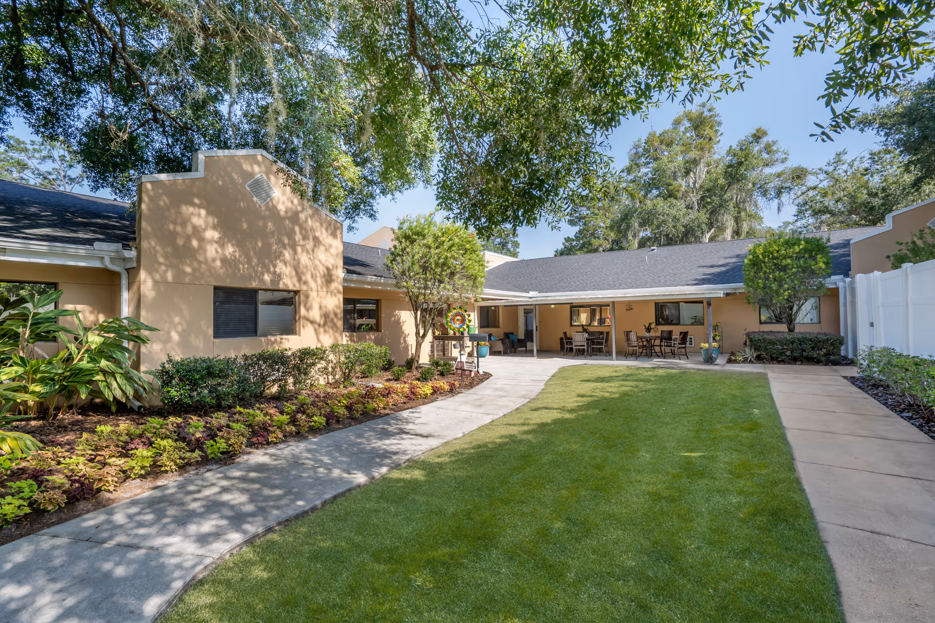 Outdoor courtyard area at Brookdale Paddock Hills featuring a well-maintained lawn, paved walkways, landscaped plants and shrubs, and a covered patio with tables and chairs under a clear blue sky.