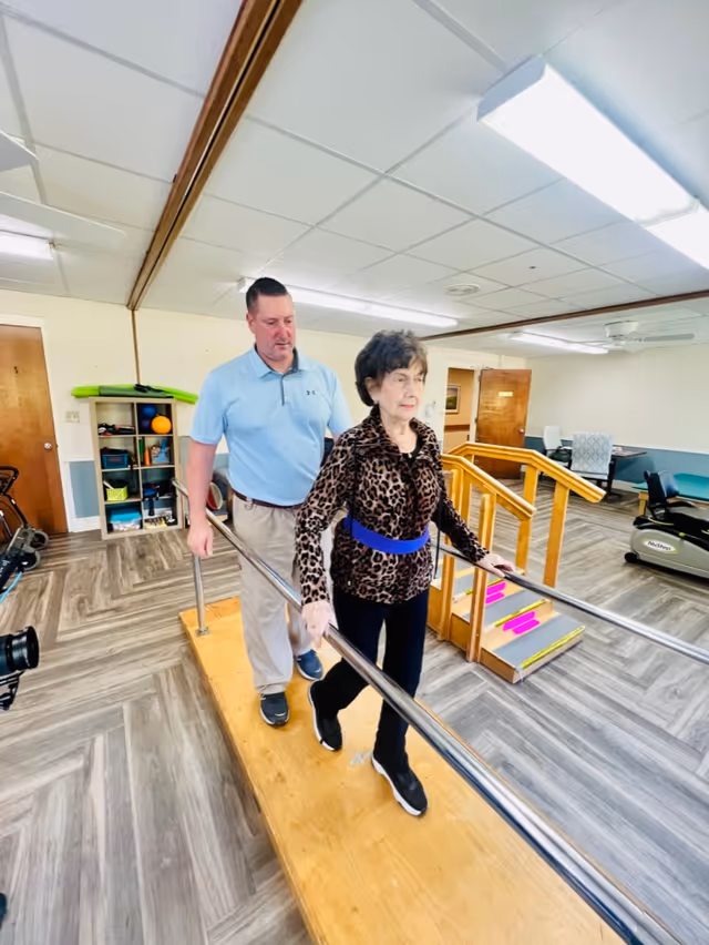 An elderly woman wearing a leopard print jacket and black pants walks on a wooden balance beam with handrails in a rehabilitation facility, assisted by a man in a light blue polo shirt and beige pants. The room has exercise equipment, a small set of stairs with colorful markings, and a shelf with various items in the background.