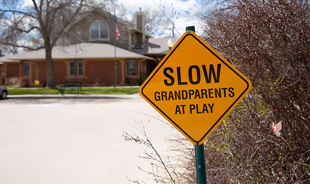A yellow diamond-shaped road sign that reads 'SLOW GRANDPARENTS AT PLAY' positioned near a bush with a residential building and trees in the background under a partly cloudy sky.