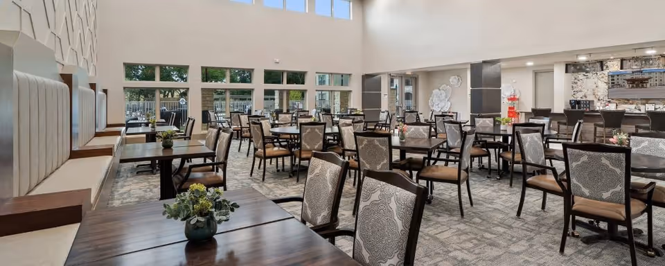 Spacious dining room with multiple tables and chairs arranged neatly. The room features large windows allowing natural light to fill the space, cushioned bench seating along one wall, decorative wall art, and small flower arrangements on the tables.