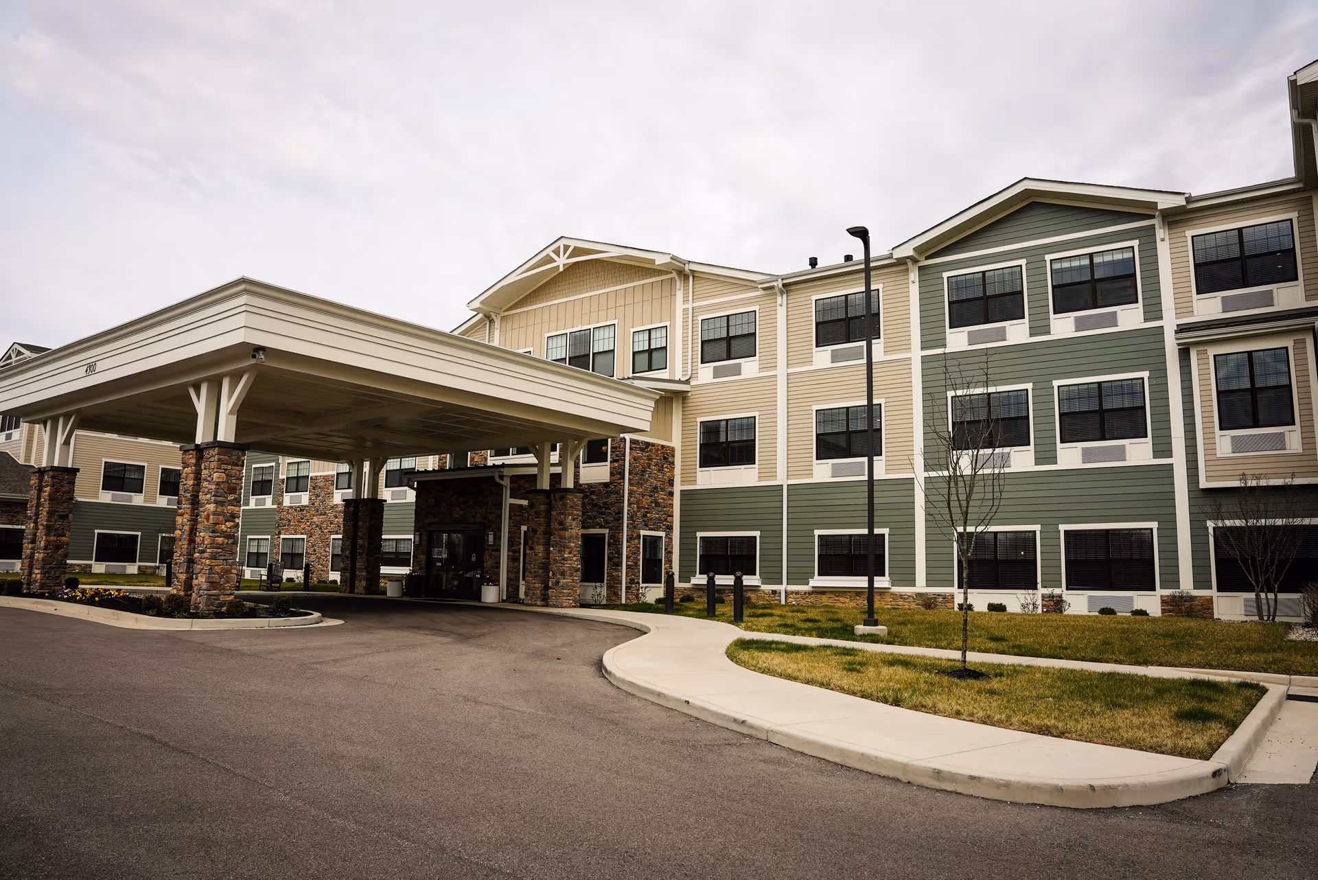 Exterior view of Traditions of Columbus senior living facility showing a three-story building with beige and green siding, multiple windows, and a covered entrance supported by stone pillars. There is a driveway and a sidewalk with some landscaping including grass and small trees.