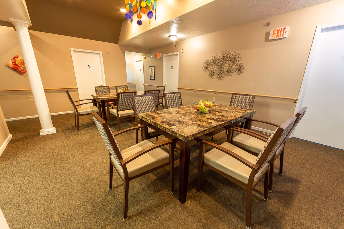 A dining area with two square tables surrounded by chairs, wall art, and an exit sign in a senior living facility.