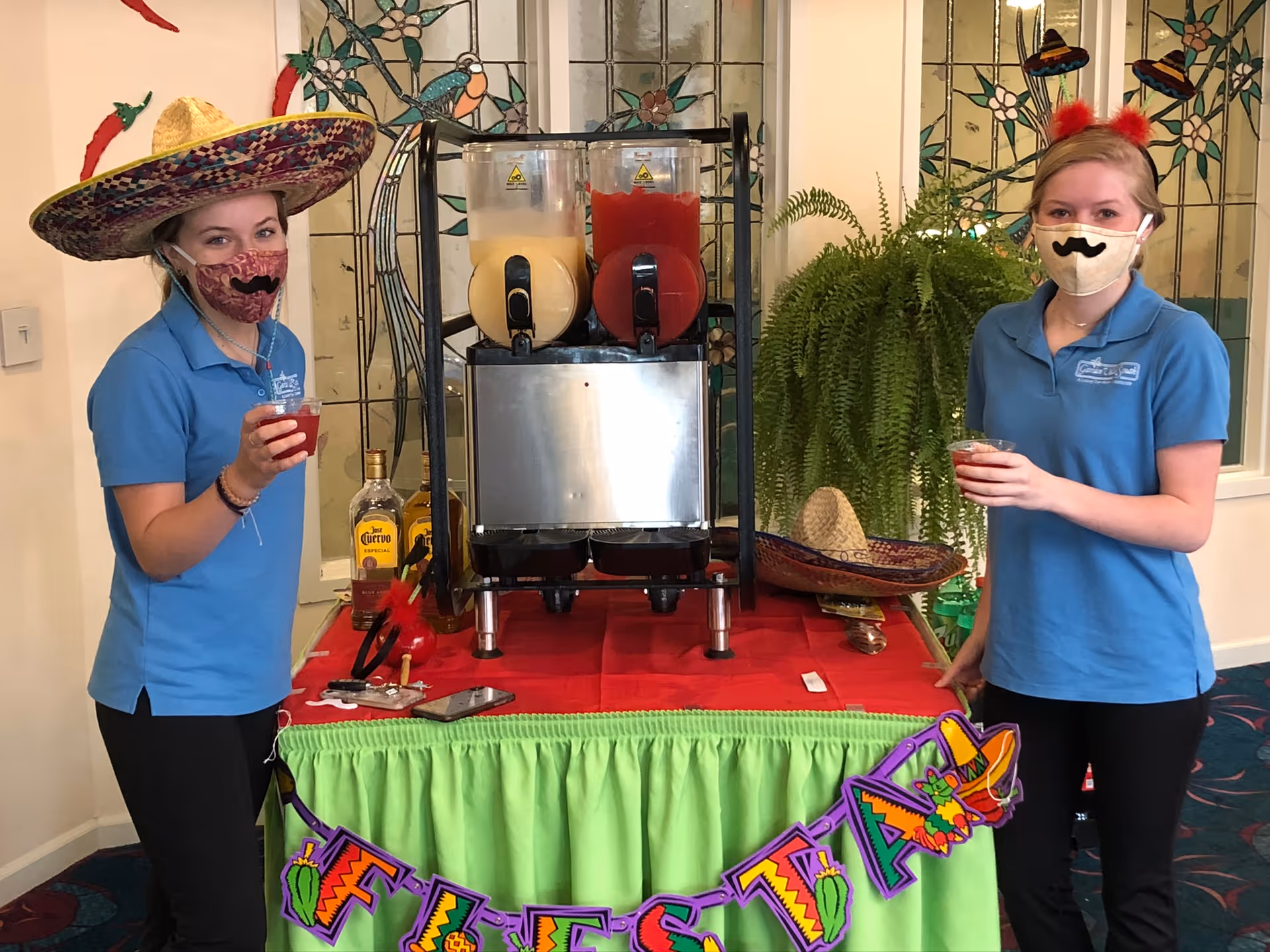Two women wearing blue polo shirts and festive masks stand on either side of a table decorated with a green skirt and a colorful Fiesta banner. One woman wears a large sombrero, and the other has a small sombrero headband. On the table are two beverage dispensers with yellow and red drinks, a bottle of Jose Cuervo tequila, and some small items. Behind them is a stained glass window and a large green fern plant.