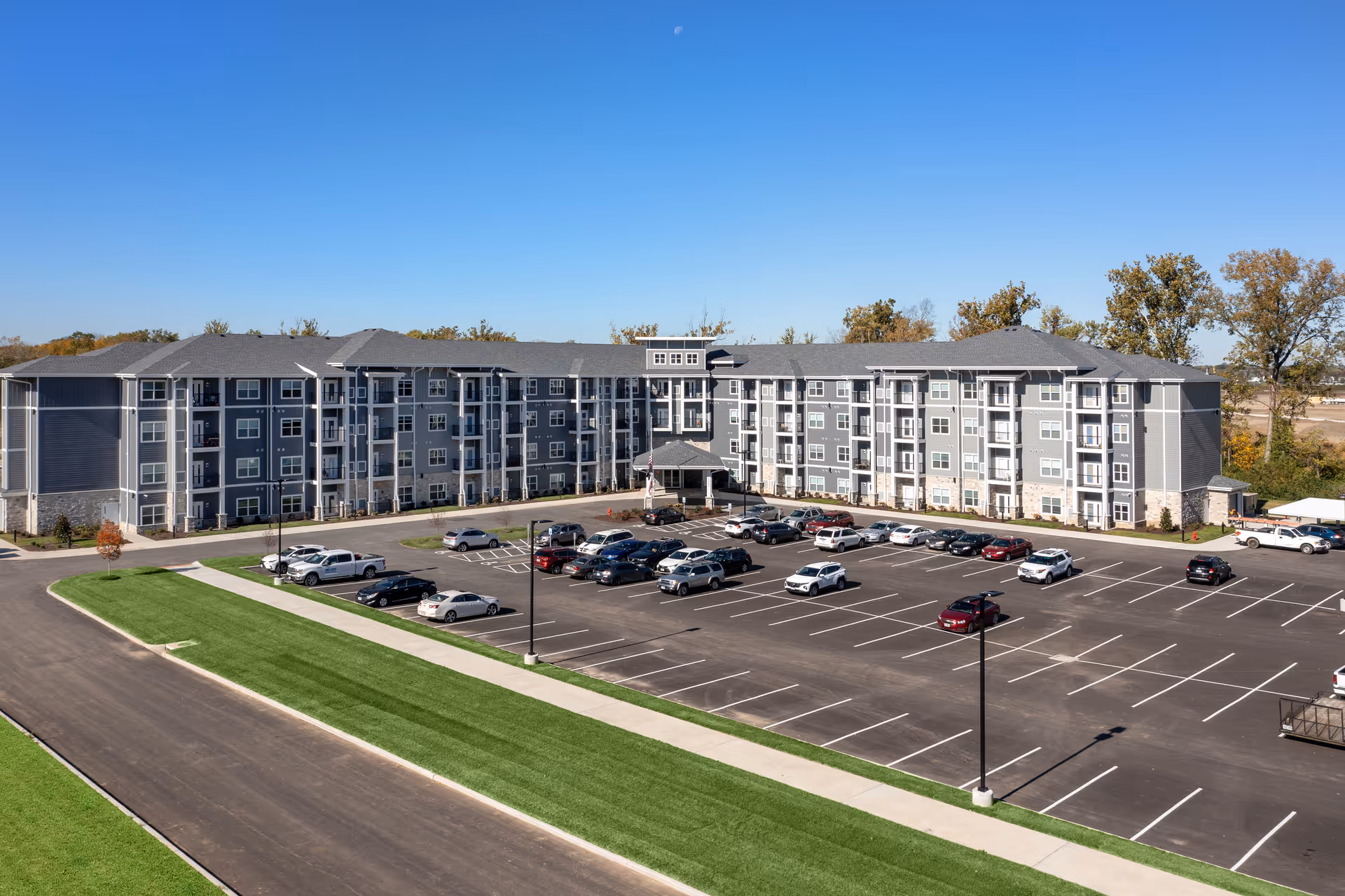 Front exterior view of a large four-story gray senior living apartment building with balconies and a mostly empty parking lot under a clear blue sky.
