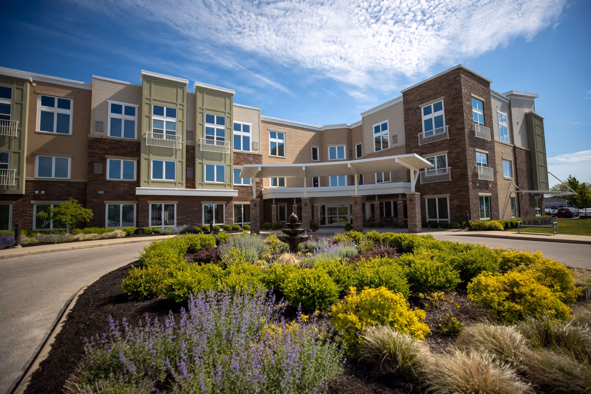 Exterior view of a three-story senior living facility building with large windows, a covered entrance, and landscaped garden with green shrubs and purple flowers in the foreground under a partly cloudy blue sky.