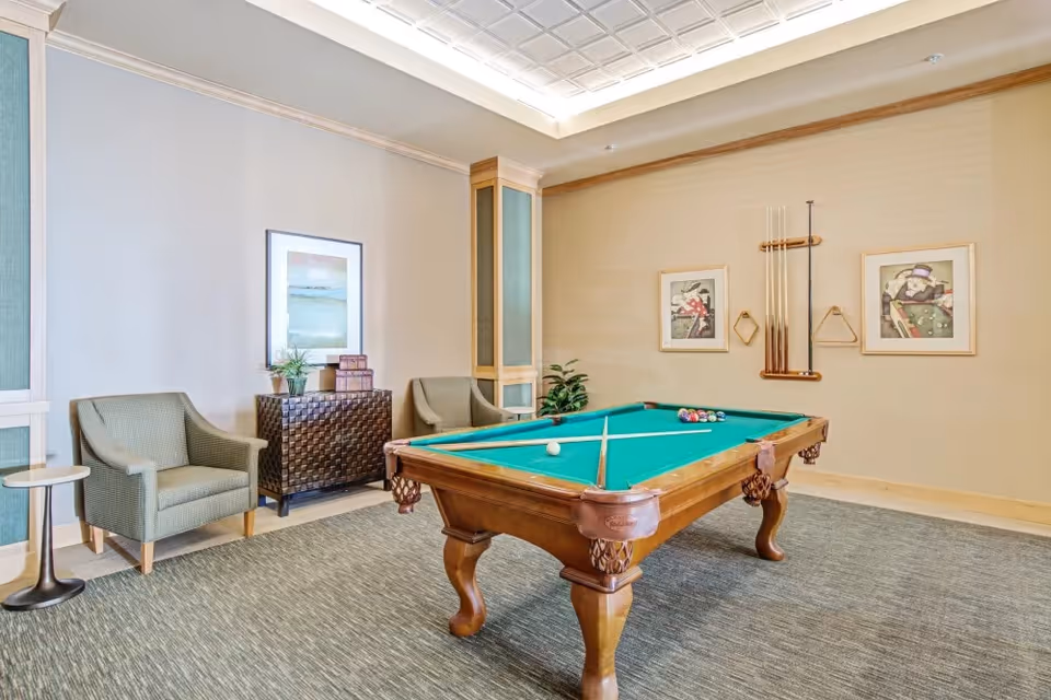 A well-lit recreational room featuring a wooden pool table with green felt, pool cues mounted on the wall, two framed pictures, two armchairs, a small side table, a decorative cabinet with plants and books, and a carpeted floor.
