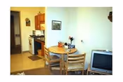 Interior view of a small apartment with a round dining table and chairs, a television, and a kitchenette with stove and cabinets.