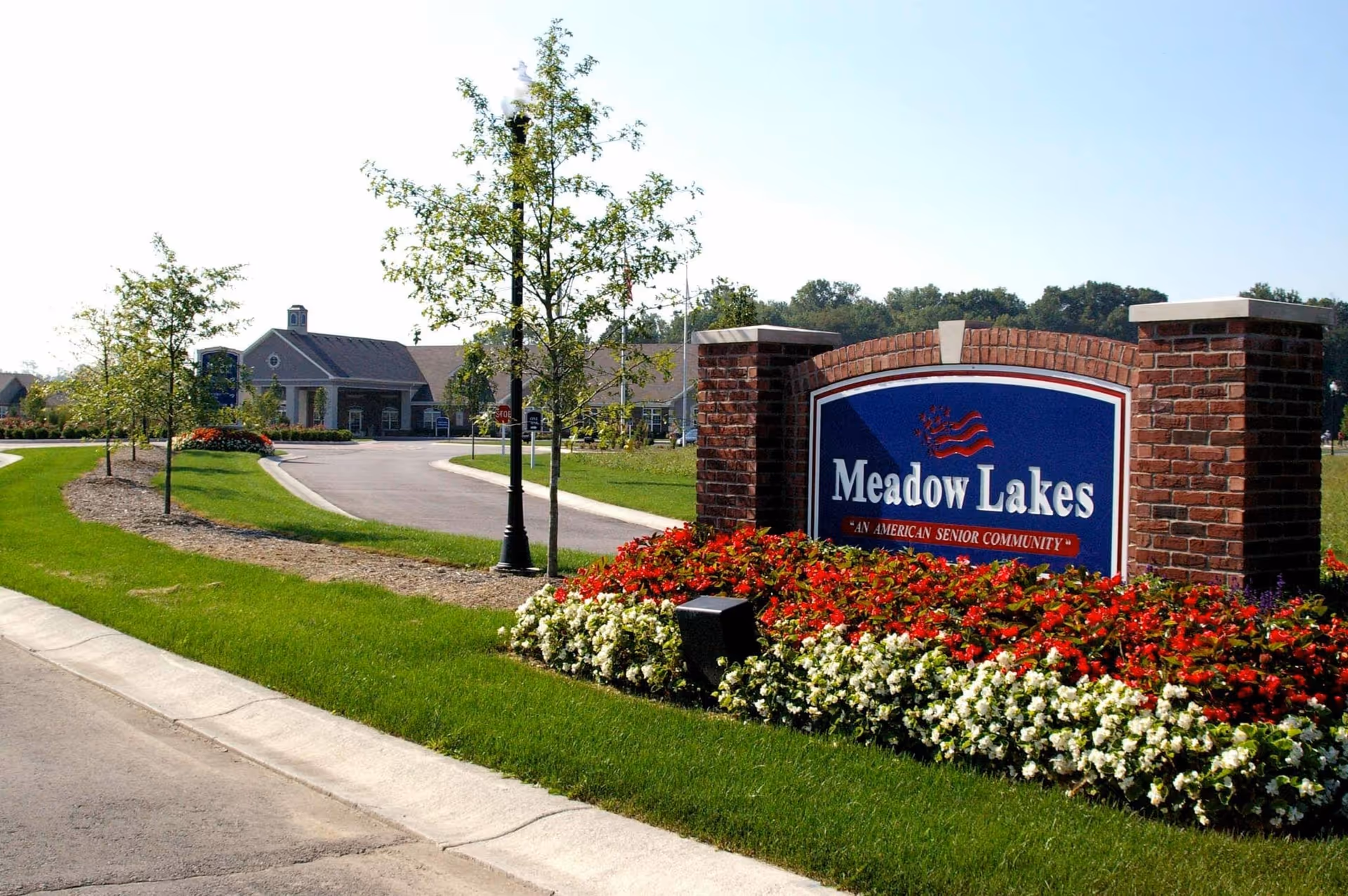 Entrance to Meadow Lakes senior community with a large brick sign surrounded by red and white flowers, a curved driveway, young trees, and a building in the background under a clear sky.
