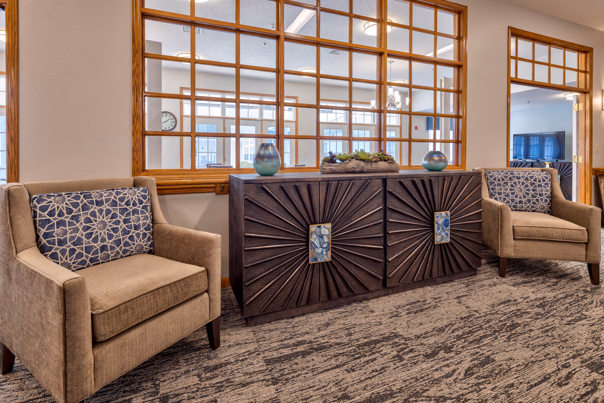 A cozy seating area with two beige armchairs featuring blue patterned cushions, positioned on either side of a dark wooden cabinet with a sunburst design on the doors. The cabinet is decorated with two blue vases and a planter with succulents. Behind the seating area is a large window with wooden frames, allowing a view into a bright room with large windows and a clock on the wall.