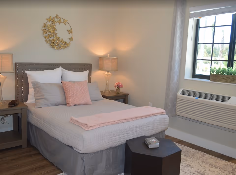 A neatly made bed with a gray quilt, white and gray pillows, and a pink throw blanket in a bedroom. There are two bedside tables with lamps on each side of the bed. A decorative gold wall art piece hangs above the headboard. A window with a white curtain and a window air conditioning unit is on the right side of the room. A small black hexagonal table with a book on it is at the foot of the bed.