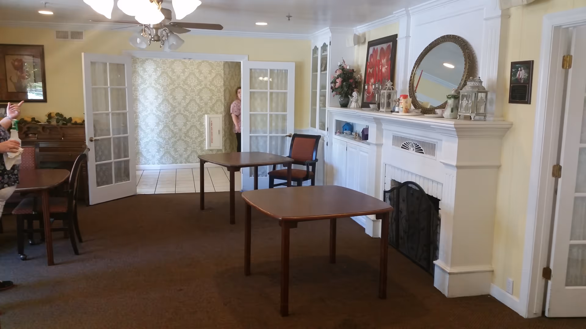 Communal sitting room with two wooden tables and chairs, a decorated white fireplace mantel, piano, and open glass French doors.