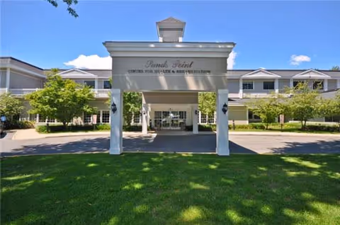 Front exterior view of Sands Point Center for Nursing & Rehabilitation building with a covered entrance, green lawn, trees, and a clear blue sky.
