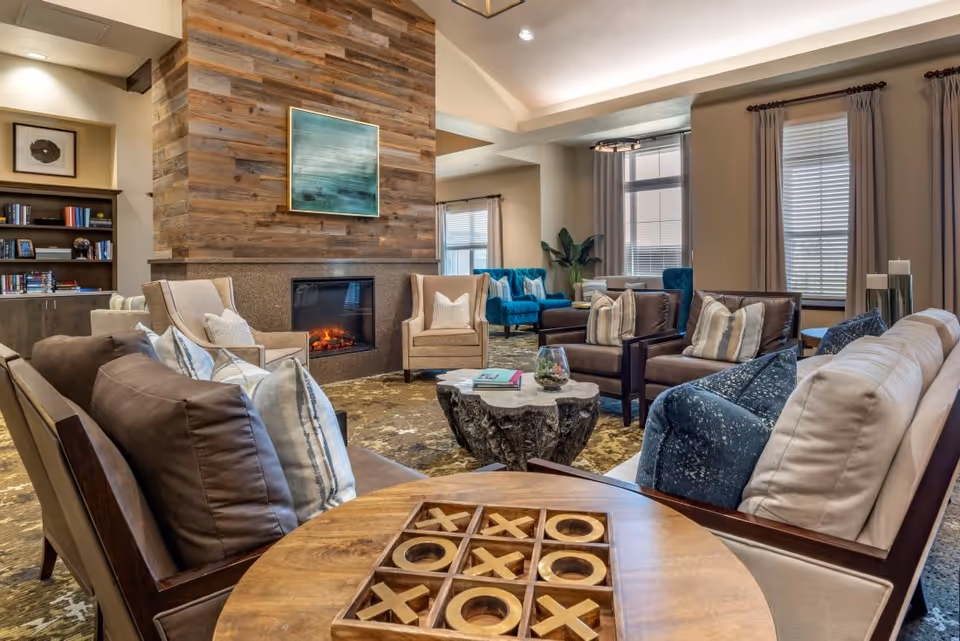 A cozy living room area in Ansel Park Assisted Living & Memory Care featuring a wooden tic-tac-toe game on a round table in the foreground, multiple cushioned chairs and sofas arranged around a rustic wooden coffee table, and a fireplace with a wooden accent wall and a framed abstract painting above it. Large windows with curtains allow natural light into the room.