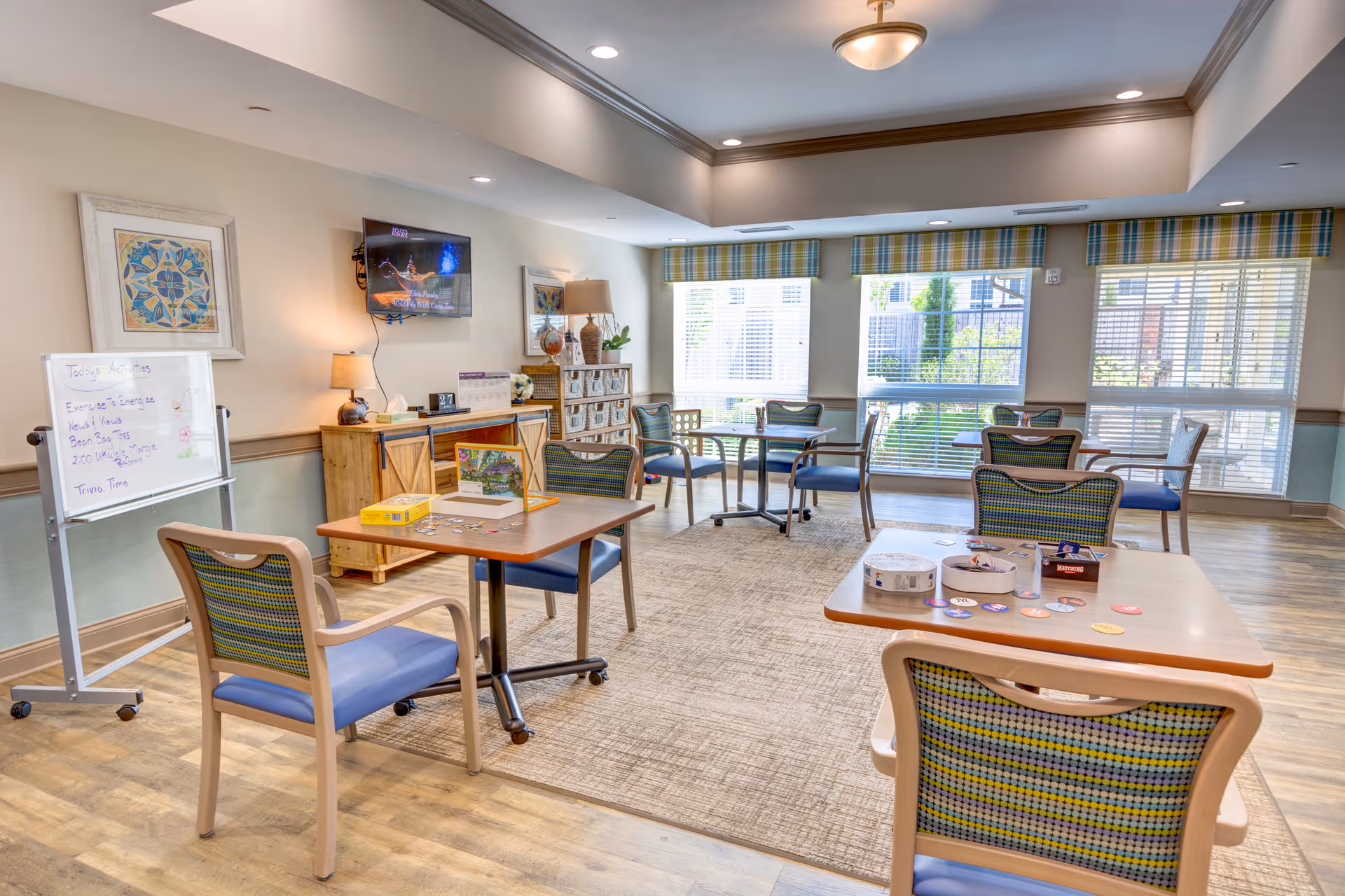 A bright and spacious activity room in a senior living facility with several tables and chairs arranged for group activities. A whiteboard with a list of activities stands on the left side, and a TV is mounted on the wall above a wooden cabinet. Large windows with blinds and plaid valances let in natural light, and the room has a cozy, welcoming atmosphere.