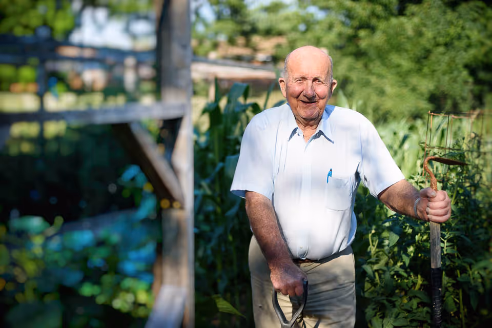 An elderly man standing outdoors in a garden holding a gardening tool, smiling and wearing a white short-sleeve shirt and beige pants, with green plants and wooden structures in the background.